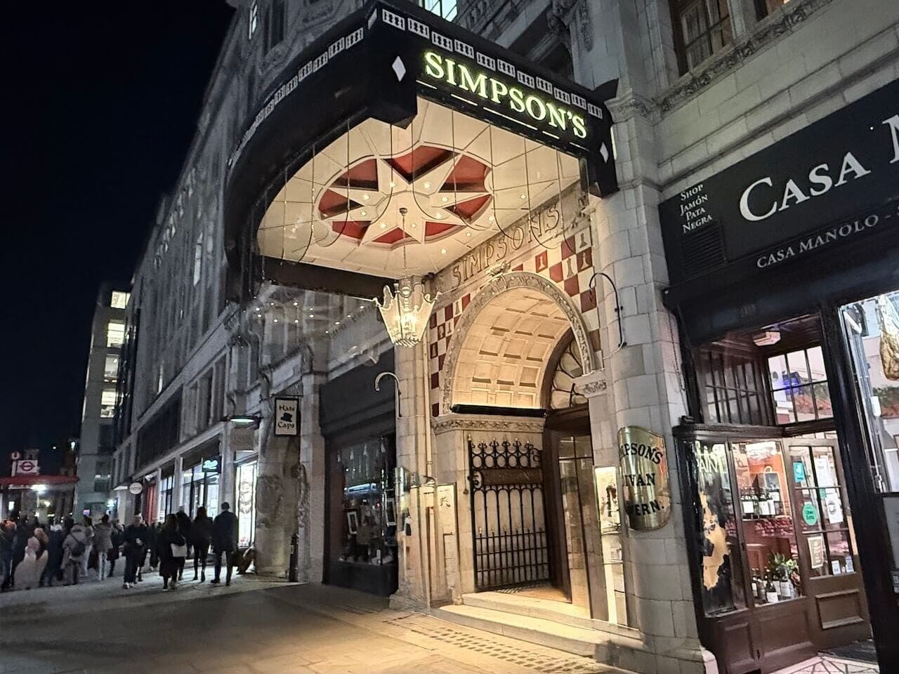 The illuminated entrance to Simpson's in the Strand on a London street at night.